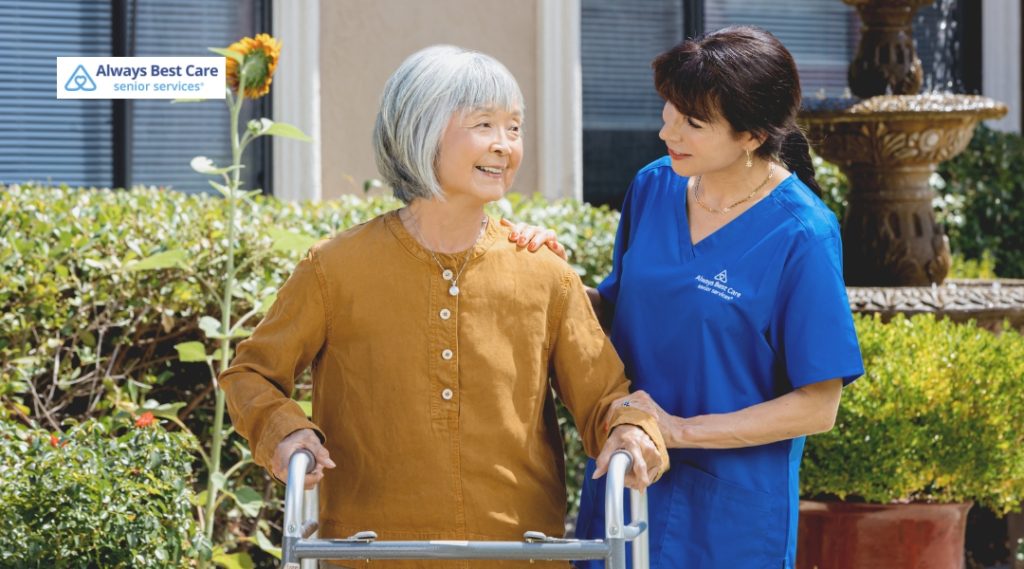 A senior woman using a walker outside, supported by a caregiver in a blue uniform, conveying compassionate, hands-on assistance and outdoor mobility support.