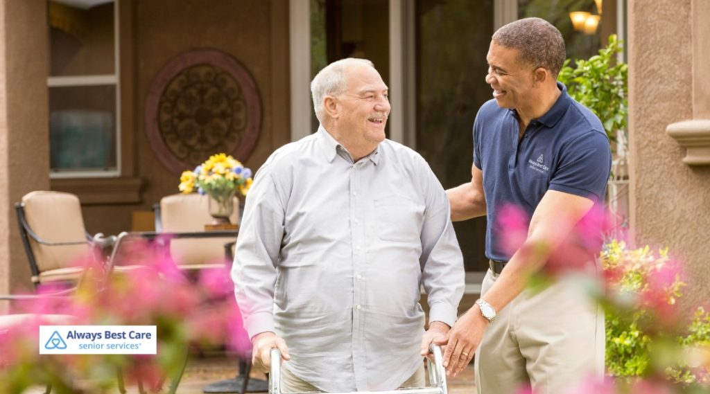 A caregiver in a green uniform assists a senior man while walking outside. The caregiver holds his arm gently, providing support as they walk through a park or garden setting, with blooming flowers around them.