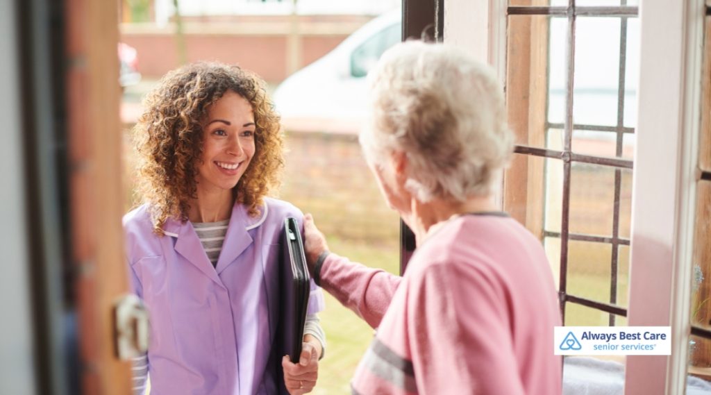 A smiling caregiver in a purple uniform greets a senior woman at the door, offering a compassionate, welcoming moment.