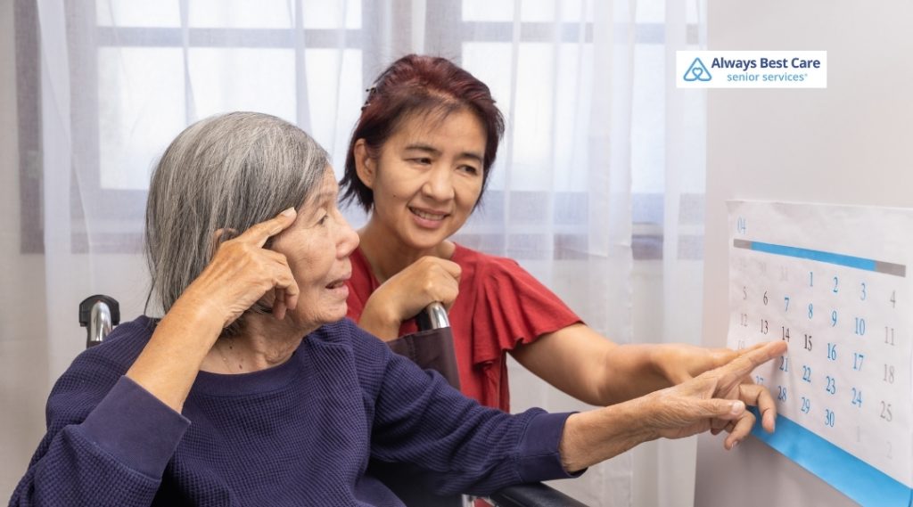 A younger woman points to a calendar on the wall while assisting an older woman in a wheelchair who appears to be experiencing memory challenges. The Always Best Care logo is in the top right corner.
