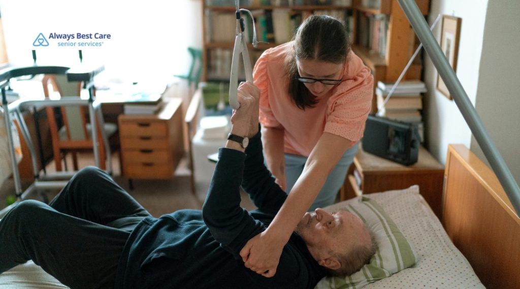 A caregiver uses a patient lift to assist a senior in moving from a bed while providing supportive care in a residential setting.