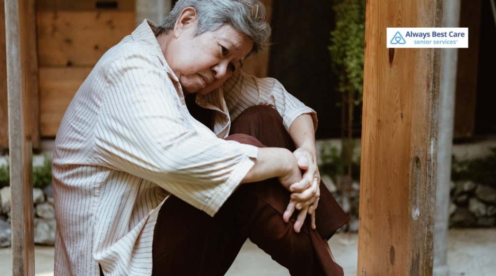Senior woman sitting curled up against a wooden post, appearing sad or withdrawn, reflecting isolation. Always Best Care logo in the corner.