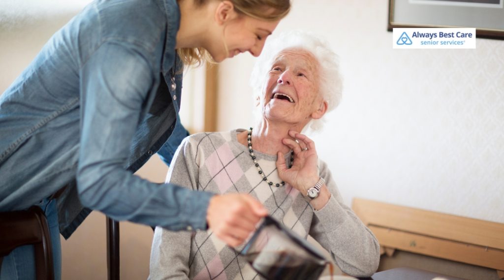 Smiling elderly woman sitting at a table, looking up joyfully at a caregiver pouring her a drink. Always Best Care logo in the corner.