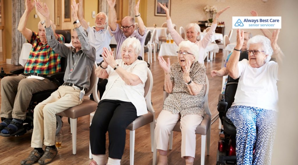 Group of seniors sitting in chairs, laughing and raising their hands in a cheerful group activity session. Always Best Care logo in the corner.