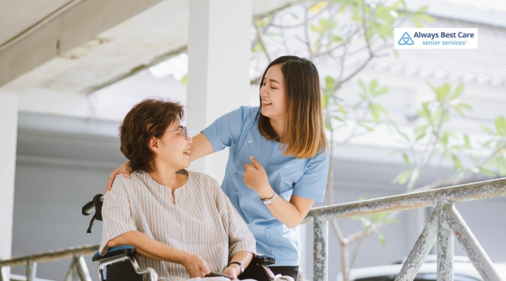 Caregiver in blue scrubs shares a joyful moment with a senior woman in a wheelchair outdoors, reflecting compassionate in-home support.