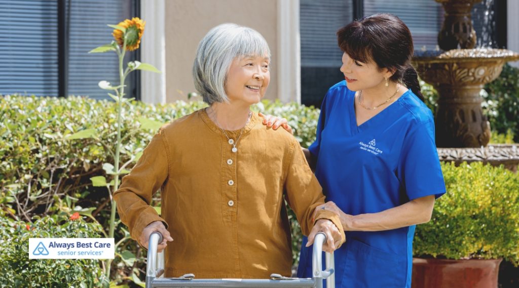 Smiling senior woman using a walker outdoors with the assistance of a caregiver in blue scrubs, demonstrating mobility support and companionship.