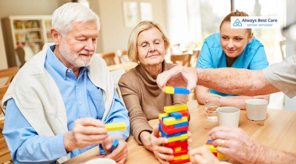 Seniors sit at a table playing a colorful stacking block game, while a caregiver in blue scrubs observes and smiles.
