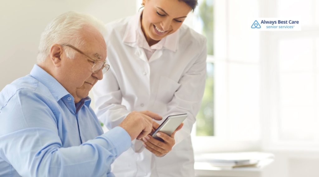 A smiling caregiver in a white coat assists a senior man using a smartphone, guiding him with a finger gesture.