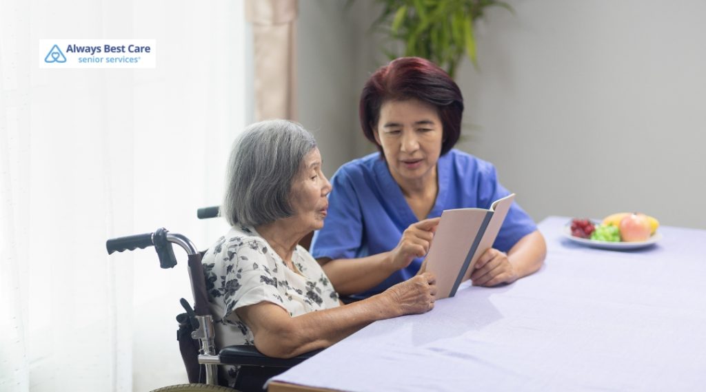 A caregiver in blue scrubs reads a book with a senior woman in a wheelchair at a table with fresh fruit in the background.