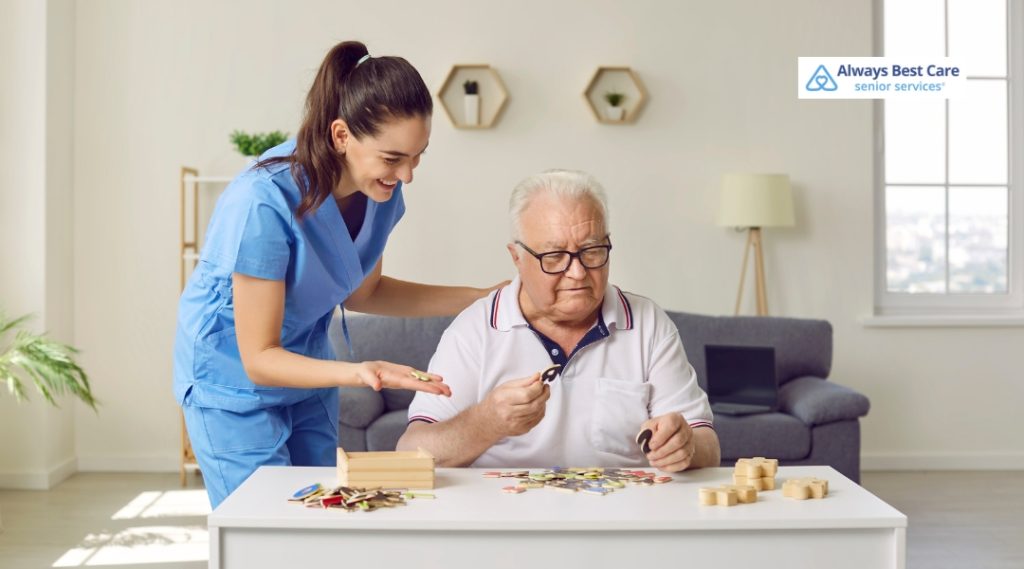 A female caregiver in blue scrubs smiles and assists an elderly man with glasses as they work on a colorful puzzle together at a white table in a bright living room.