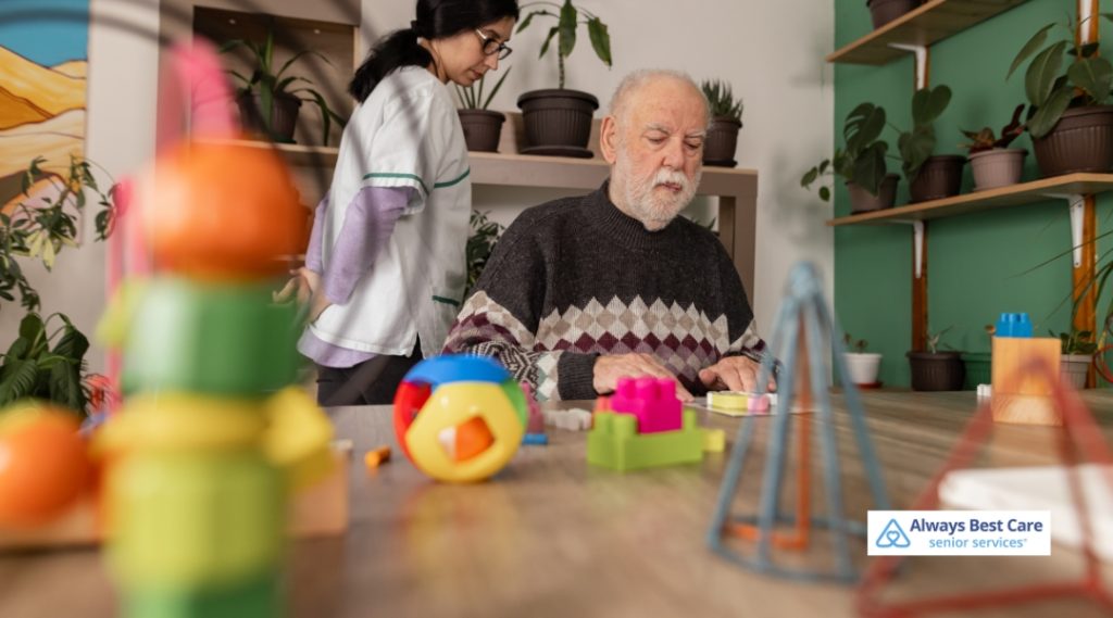 An elderly man with a white beard focuses on colorful building blocks at a table, while a caregiver stands nearby in a room filled with potted plants and toys.

