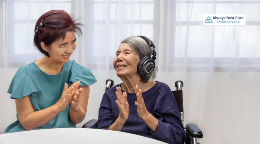 A smiling elderly woman in a wheelchair wears large headphones and claps along with music, accompanied by a woman in a green top who is also clapping and smiling.