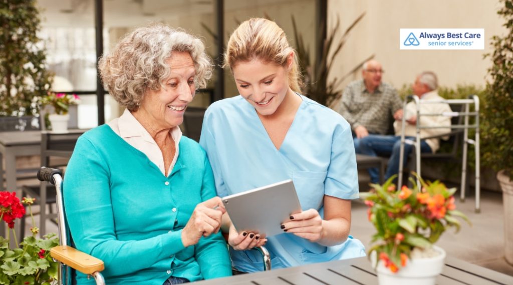 A smiling caregiver in light blue scrubs shows a tablet to a senior woman in a wheelchair, both enjoying the moment in an outdoor patio area.