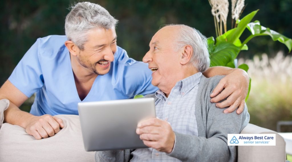  A caregiver in blue scrubs shares a laugh with a senior man while holding a tablet, creating a warm and friendly connection.