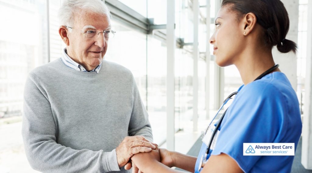 A caregiver in blue scrubs gently holds the hand of a senior man in glasses, offering reassurance in a bright, modern setting.