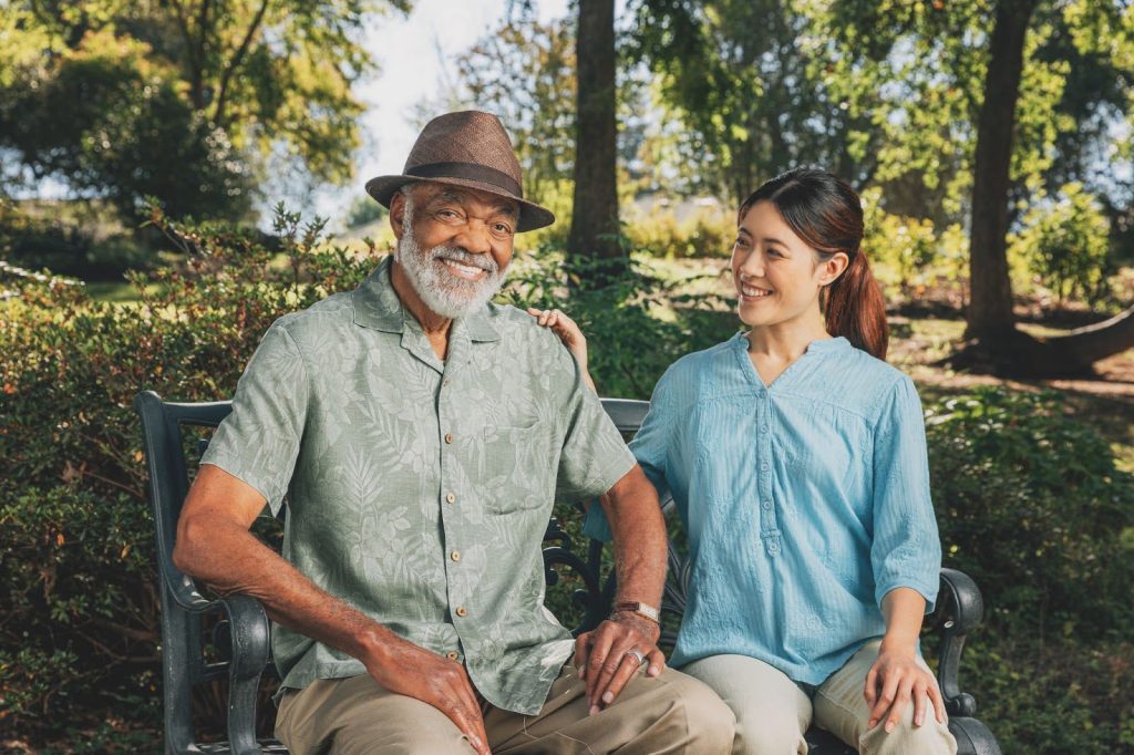 A smiling senior man wearing a brown hat sits on a park bench with a young female caregiver in a light blue blouse beside him. They enjoy a sunny day surrounded by trees and greenery, sharing a friendly conversation.