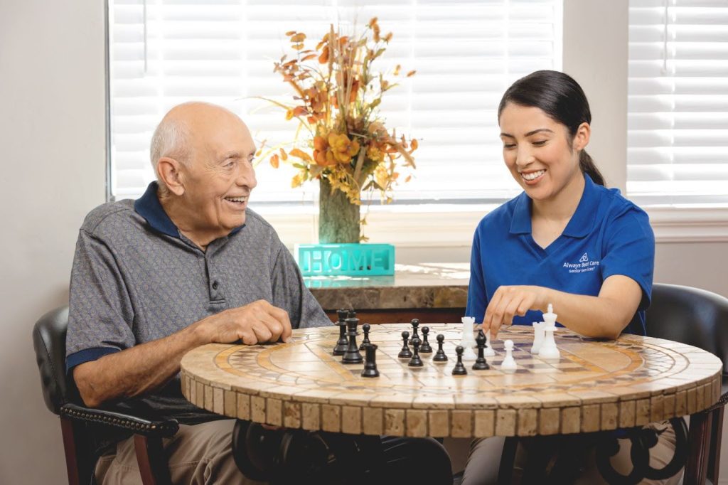 A cheerful senior man plays chess with a caregiver wearing a blue Always Best Care polo shirt. They sit at a round mosaic table in a bright indoor setting decorated with autumn flowers and a “HOME” sign.