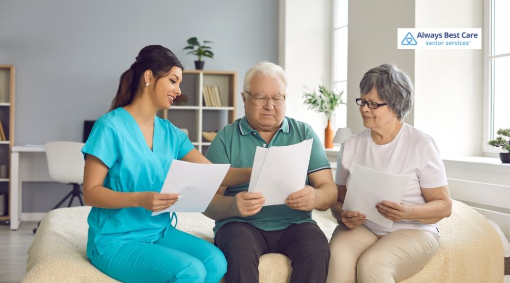 A senior couple is seated on a couch with an Always Best Care representative, reviewing paperwork. The couple appears comfortable and engaged in the discussion.