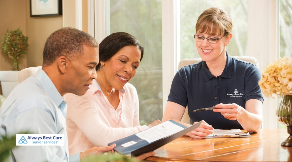 A senior couple is sitting on a couch, looking at documents with an Always Best Care nurse. The couple is reviewing information, while the nurse points to the paperwork, and they all appear to be in a positive, collaborative conversation.