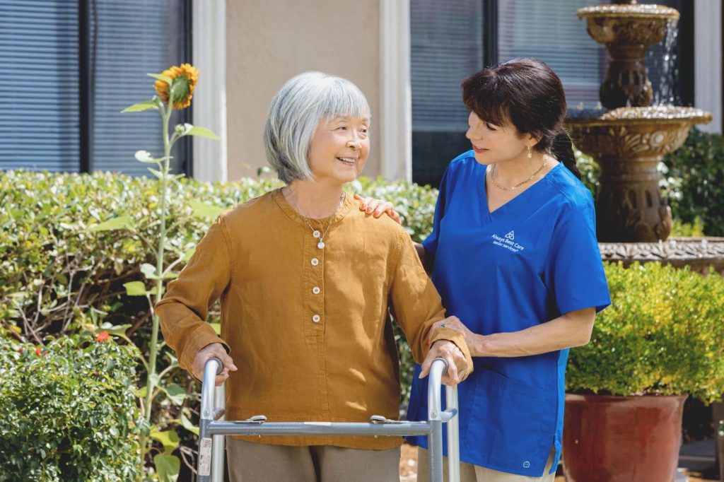 An older woman using a walker smiles as a caregiver in a blue Always Best Care scrub top offers gentle support on her shoulder. They are outdoors near a garden fountain and colorful flowers.