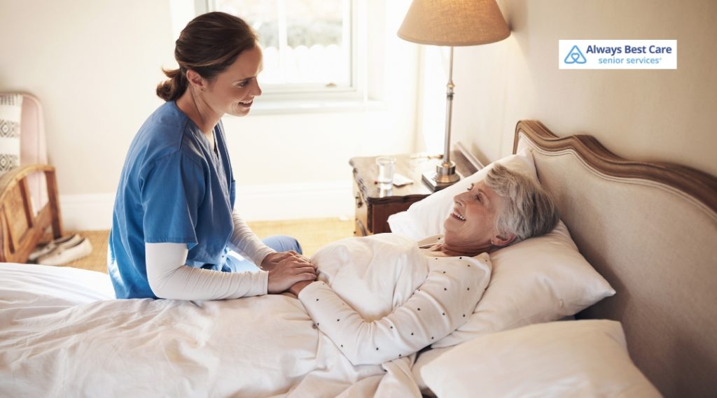 A compassionate caregiver in blue scrubs gently holds the hand of an elderly woman with gray hair, as she rests in bed. The caregiver is smiling and offering comfort in a well-lit room. The Always Best Care logo is placed in the top-right corner.