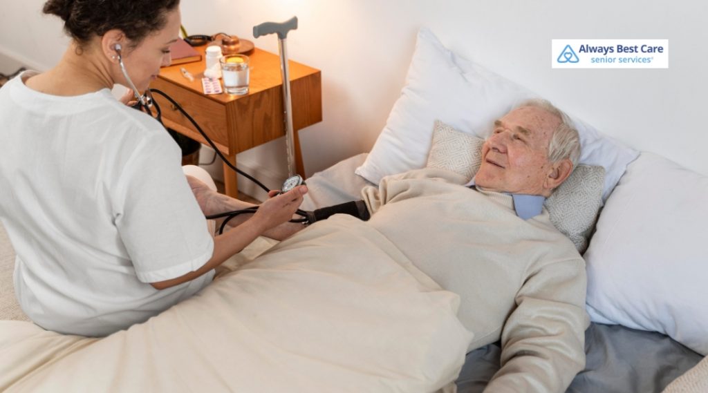 A caregiver in a white shirt uses a stethoscope to check the blood pressure of an elderly man in bed. The man is smiling and appears relaxed, while a bedside table with medication and a glass of water is visible. The Always Best Care logo is located in the top-left corner.