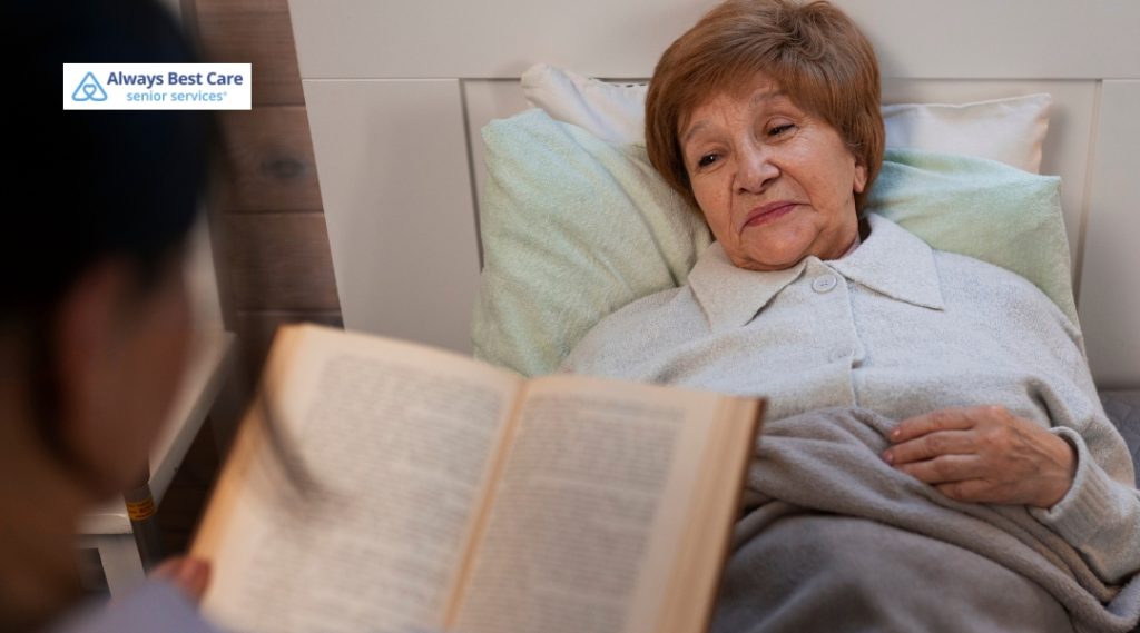 A senior woman in bed listens attentively as a caregiver reads aloud from a book. The caregiver and elderly woman are in a calm and peaceful home setting, with natural light streaming through the window. The Always Best Care logo appears in the top-left corner.
