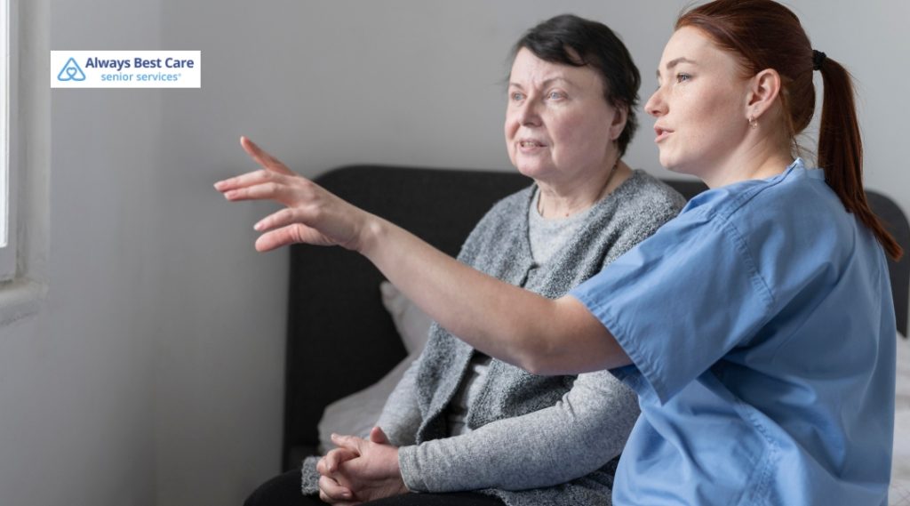 A caregiver in blue scrubs sits beside a senior woman in bed, gently talking to her and pointing towards the window, perhaps showing something outside. The room has soft lighting, creating an intimate and supportive atmosphere. The Always Best Care logo is visible in the top-left corner.