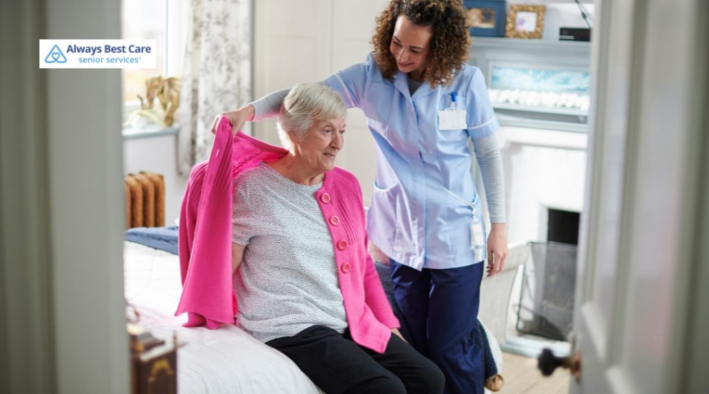 A caregiver helps a senior woman with her pink cardigan as she sits on the edge of her bed. The caregiver offers assistance with a warm smile, and the setting appears cozy and home-like. The Always Best Care logo is visible in the top-left corner.