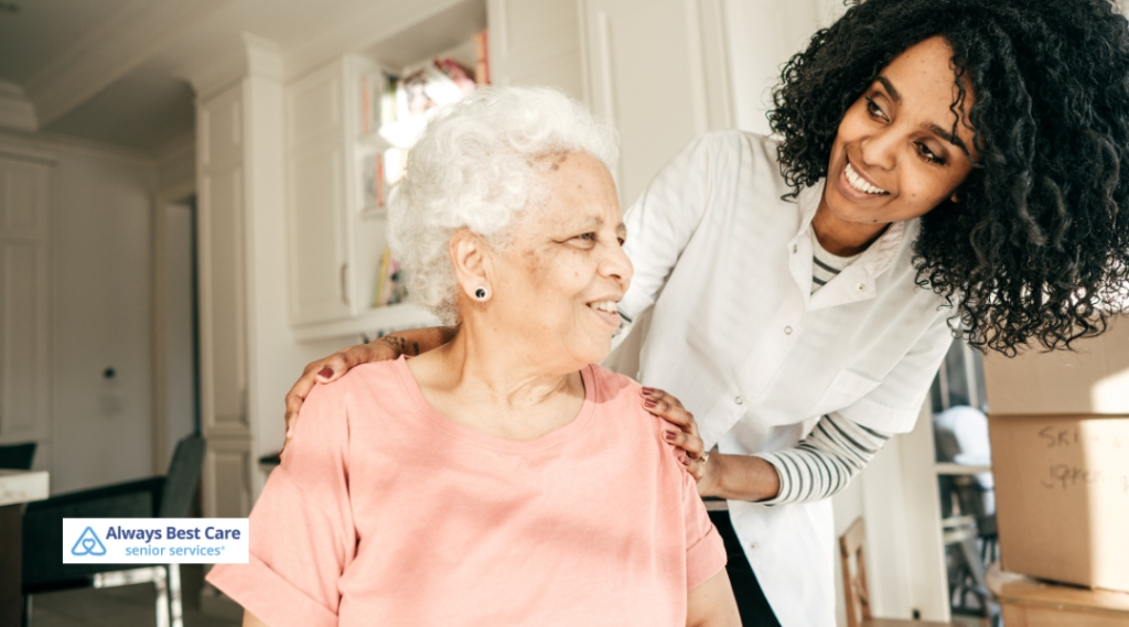 A caregiver stands behind a smiling elderly woman in a pink shirt, offering comfort and support in a bright home setting.