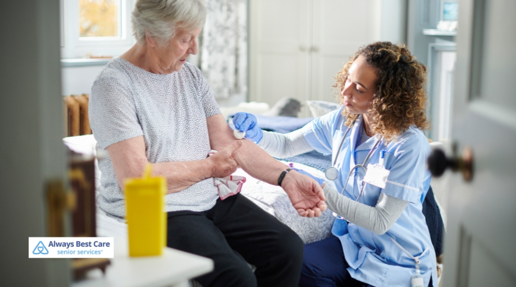 A nurse wearing blue scrubs provides medical care to an elderly woman sitting on a bed, gently holding her arm while preparing to administer treatment.