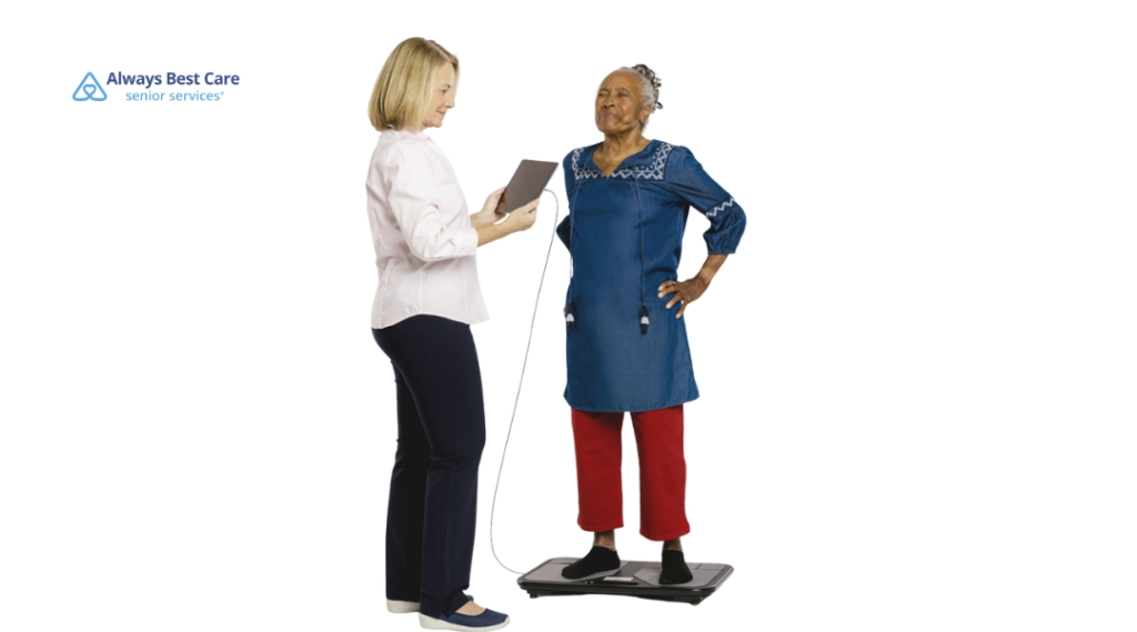 A caregiver assisting an elderly person standing on a scale, both of them smiling as the caregiver uses a tablet to monitor the person's health.