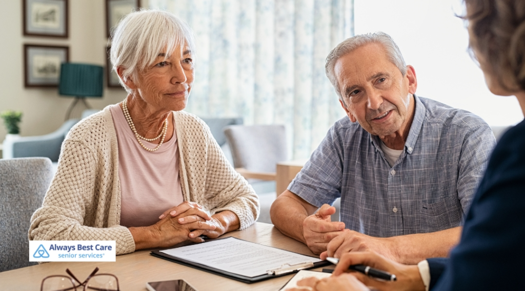 An elderly couple is seated with an Always Best Care representative, reviewing a document at a table. They are all smiling and engaged in a friendly discussion.