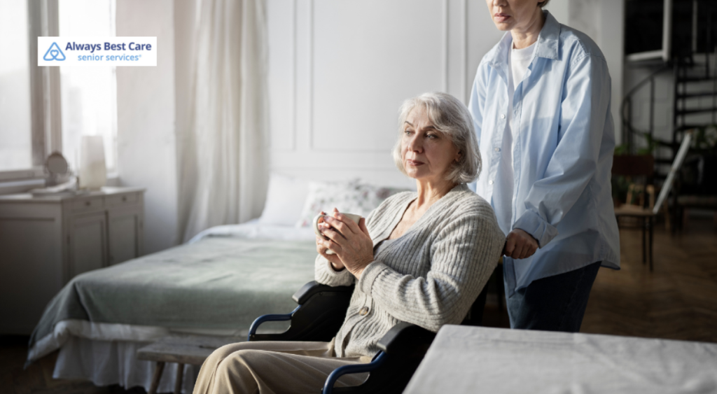 An elderly woman, with silver hair, sits in a wheelchair holding a mug, while a caregiver in a light blue shirt assists her by pushing the chair. They are inside a bright, modern room with a peaceful atmosphere, and the Always Best Care logo is visible.
