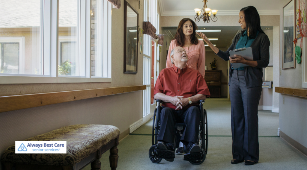 A caregiver in a blue uniform, with a clipboard, discusses care details with a senior man in a wheelchair, while a woman standing next to them listens attentively. The scene takes place in a well-lit hallway, and the Always Best Care logo is in the corner.