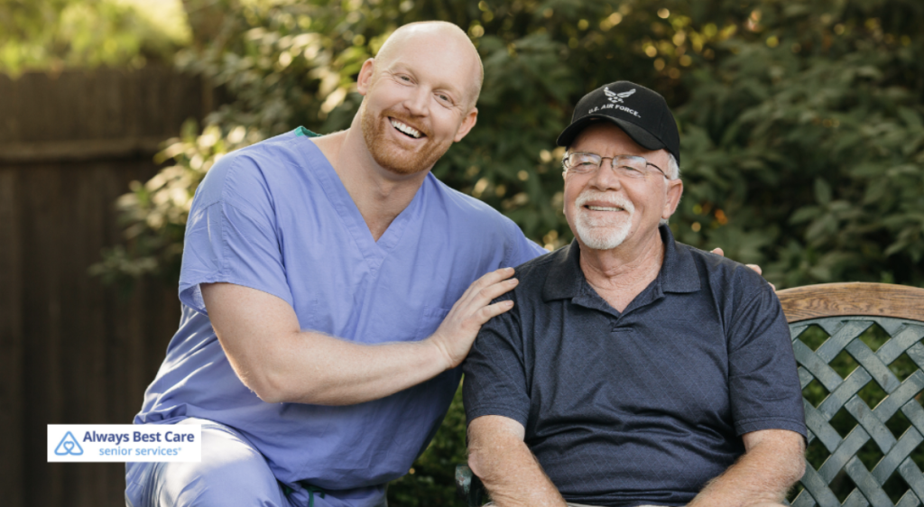 A caregiver and a senior man, both smiling, are sitting on a wooden bench outdoors in a lush, green environment. The caregiver, dressed in a blue uniform, has her arm around the man, showing warmth and companionship. The Always Best Care logo appears in the corner.