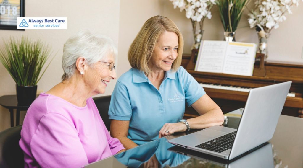 A caregiver and a senior woman are seated at a table, sharing a light moment while looking at a laptop. The caregiver wears a blue Always Best Care shirt, and the senior woman is dressed in a pink top.