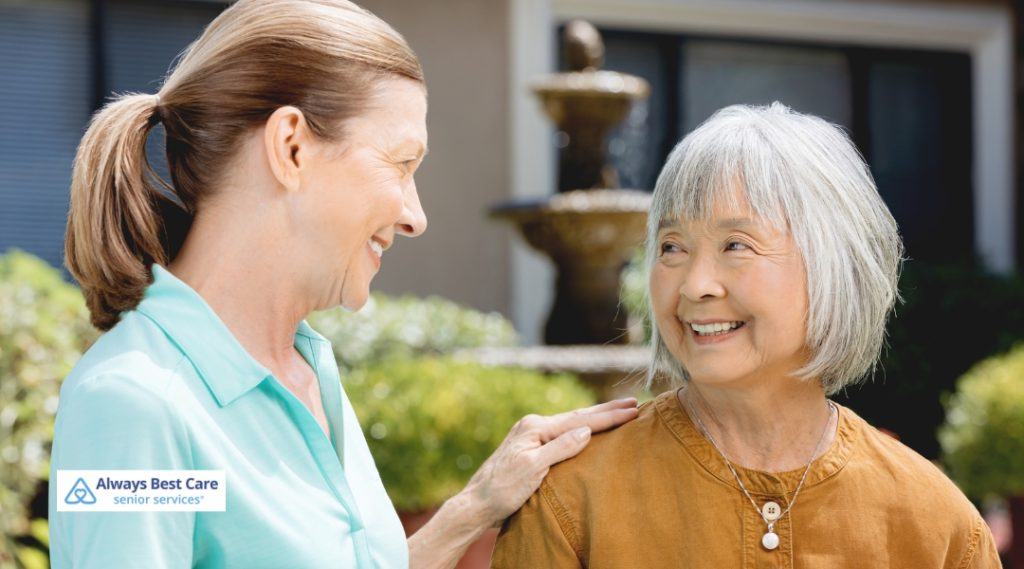 A caregiver and a senior woman share a joyful conversation outdoors, with the caregiver wearing a blue Always Best Care shirt and the senior woman wearing a brown top. They stand next to a fountain in a sunny, green garden.