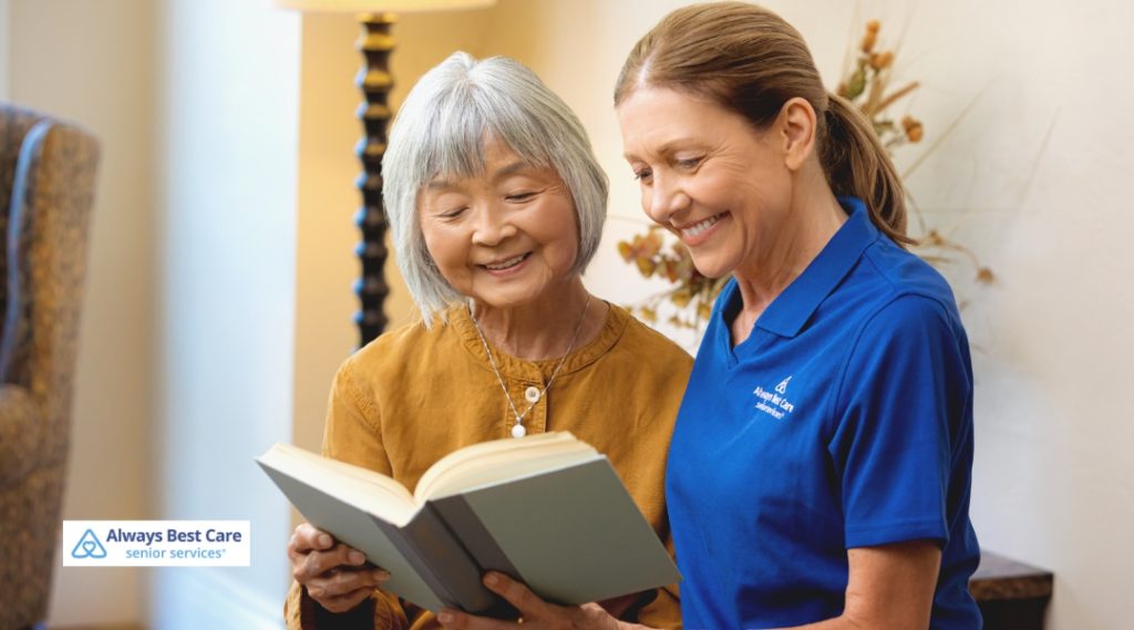 A caregiver and a senior woman are sitting together, looking at a book with a warm smile. The caregiver wears a blue Always Best Care shirt, and the senior woman is dressed in a mustard-colored top.