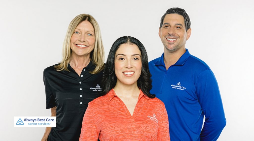 A group photo of three Always Best Care team members wearing branded shirts, smiling at the camera. The person on the left wears a black shirt, the middle person wears an orange shirt, and the person on the right wears a blue shirt.