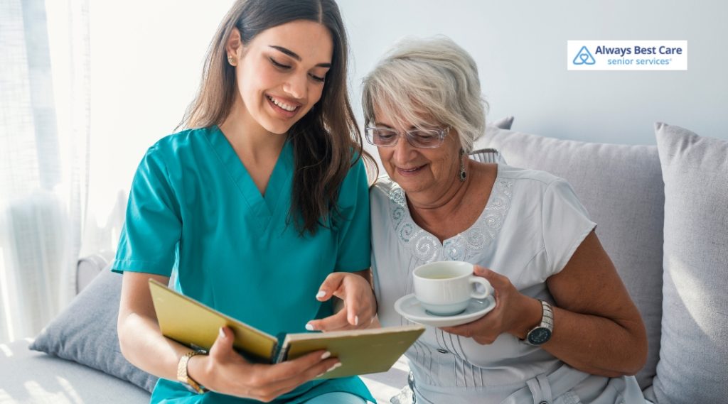 A smiling caregiver in teal scrubs reads a book with an older woman who is holding a teacup. They sit closely on a bright couch, sharing a joyful moment. The Always Best Care logo appears in the corner.