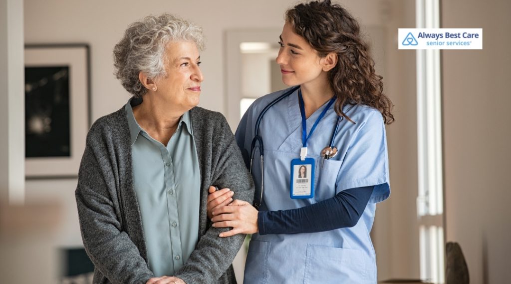 A female caregiver with a stethoscope gently supports an elderly woman walking in a home environment. They exchange a warm, trusting look, showcasing compassionate care. The Always Best Care logo is displayed.