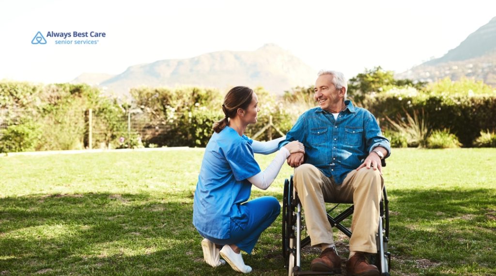 A female caregiver kneels next to an older man in a wheelchair outside on a sunny day. Both are smiling and enjoying the fresh air with scenic mountains in the background. The Always Best Care logo is present.