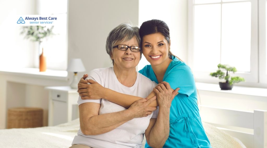 A smiling female caregiver embraces a senior woman while seated together on a bed in a sunlit room. Their close bond conveys warmth and trust. The Always Best Care logo is shown in the image.