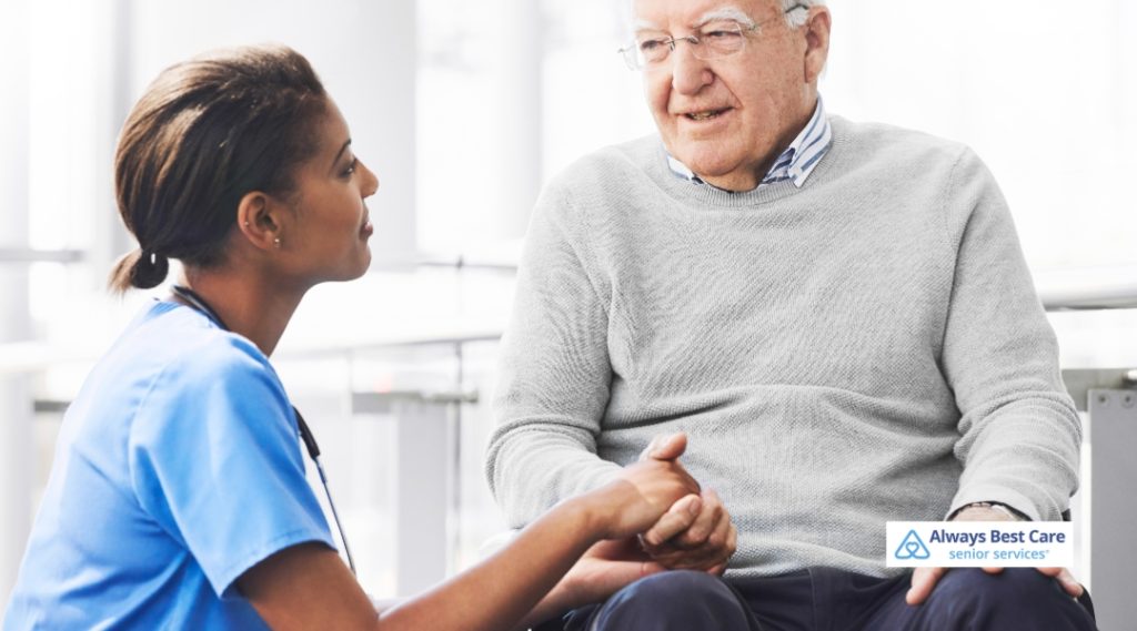 A female caregiver kneels beside an older man, gently holding his hand while they have a heartfelt conversation. The setting is modern and well-lit. The Always Best Care logo is visible.