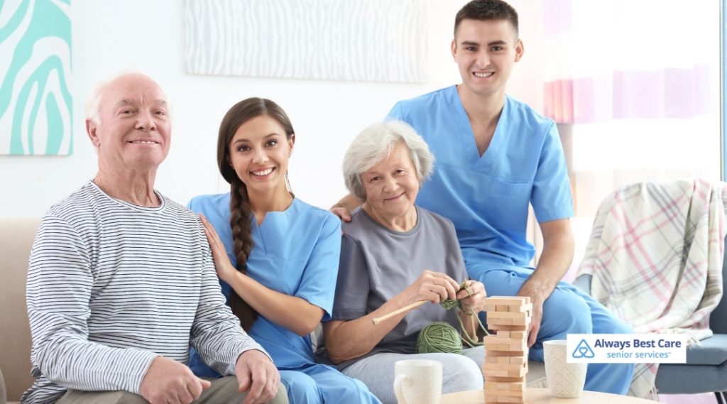 Two caregivers sit with two happy seniors during an engaging activity session. One senior knits while the group smiles at the camera. A stack of Jenga blocks and yarn are on the table, promoting companionship and cognitive stimulation. The Always Best Care logo is included.