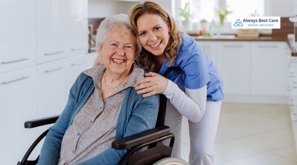 A cheerful caregiver in blue scrubs leans in to embrace an elderly woman in a wheelchair, both smiling warmly in a bright kitchen.