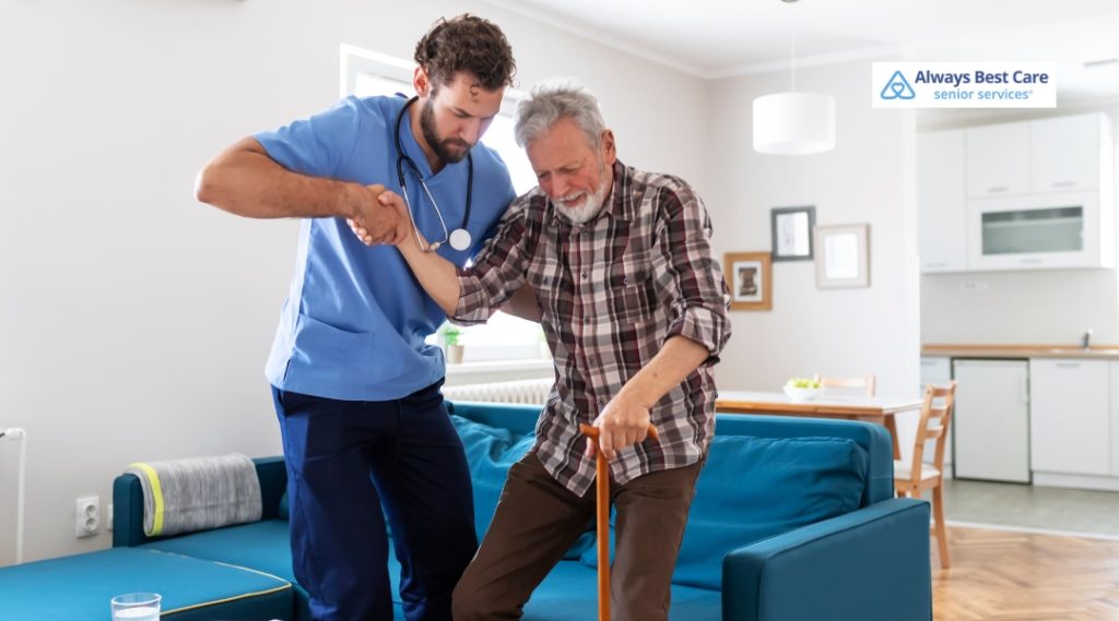 A caregiver in blue scrubs supports an elderly man with a cane, helping him stand up in a cozy living room.