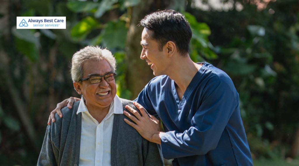 A smiling caregiver in navy scrubs walks outdoors with a senior man, sharing a lighthearted conversation.