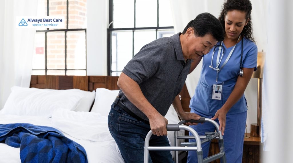 A caregiver helps an elderly man using a walker stand up safely from his bed in a bright room.